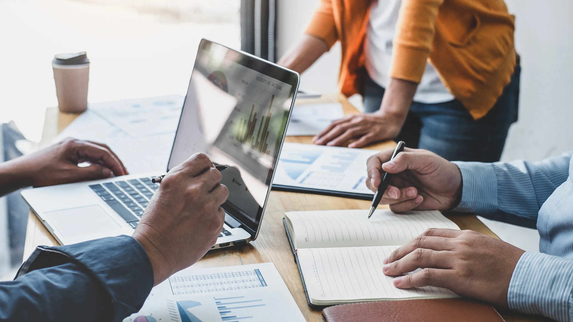 Three people collaborate at a desk, using a laptop and reviewing documents. A person takes notes, suggesting a focused, professional atmosphere.
