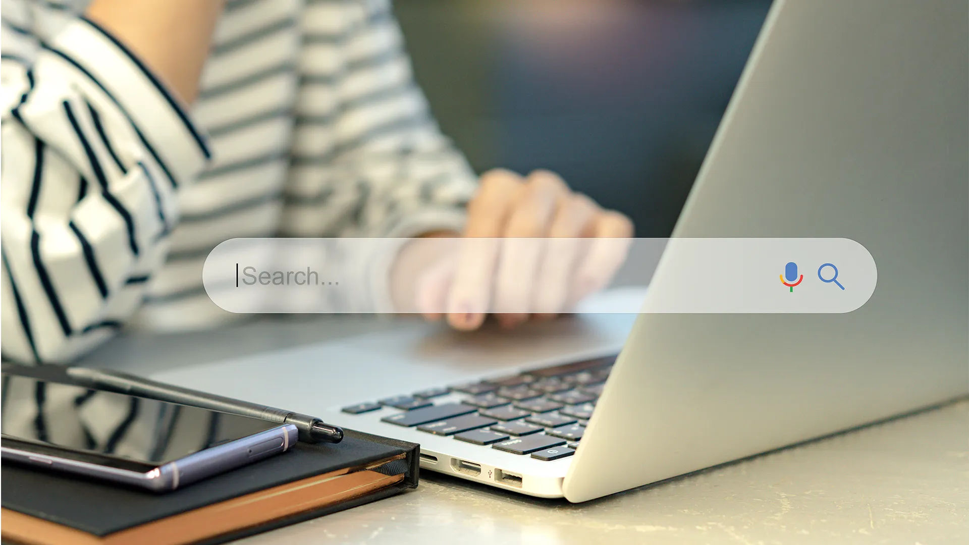 A person in a striped shirt uses a laptop, next to a notebook with a pen. A floating search bar suggests online searching. Calm, focused atmosphere.