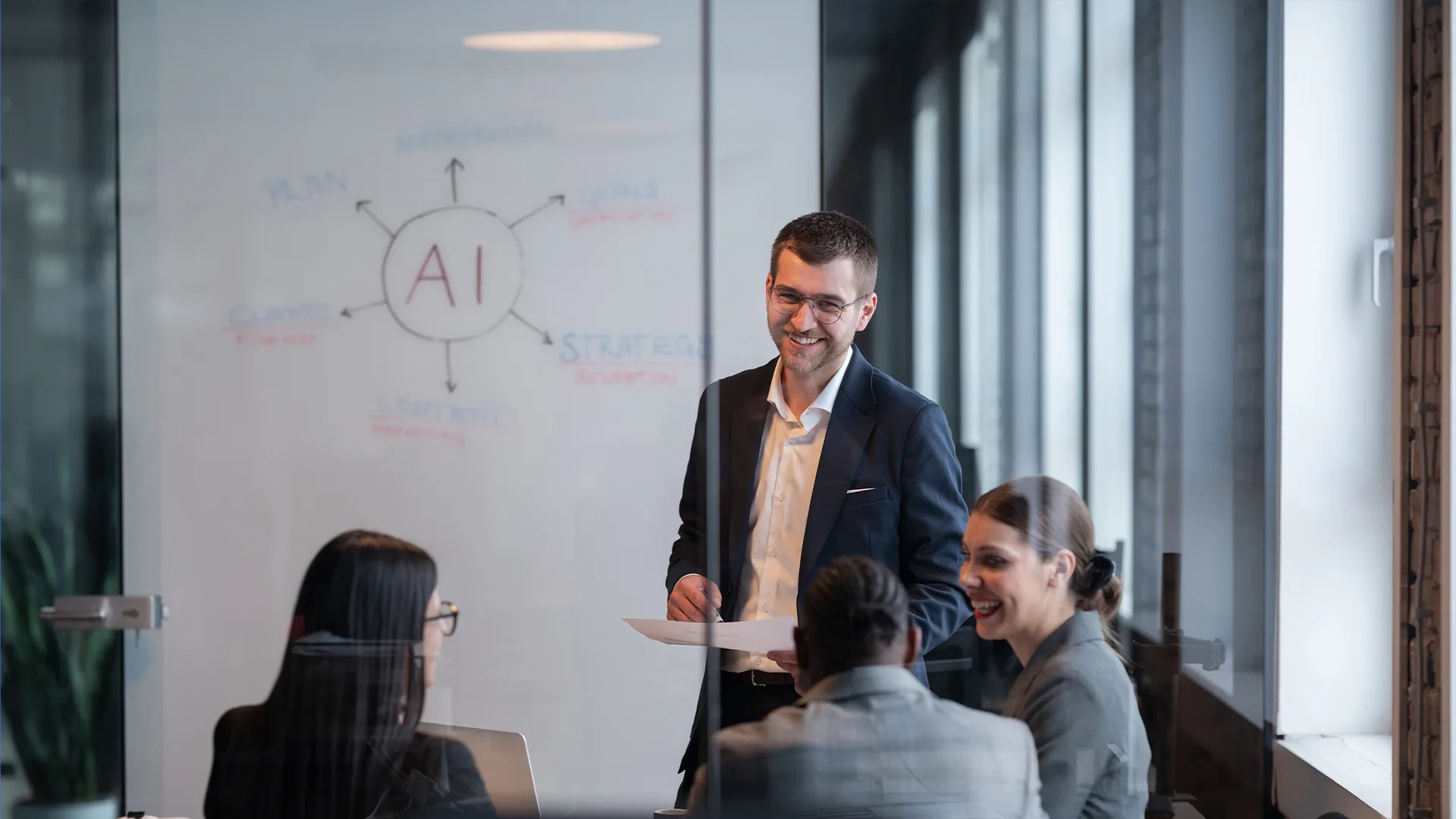 A group of business professionals in a glass-walled meeting room discuss AI strategies. A man stands, smiling and holding papers, while four seated colleagues listen attentively.