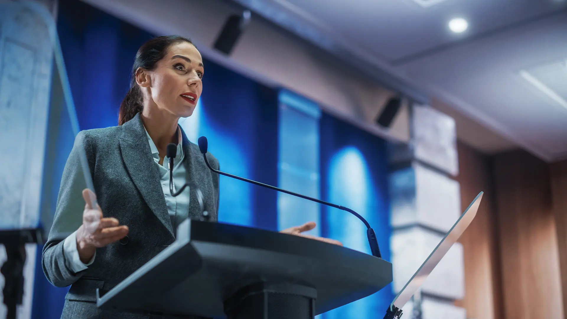 A woman in a suit speaks passionately at a podium with a microphone, set against a blue-lit background. The atmosphere is professional and focused.