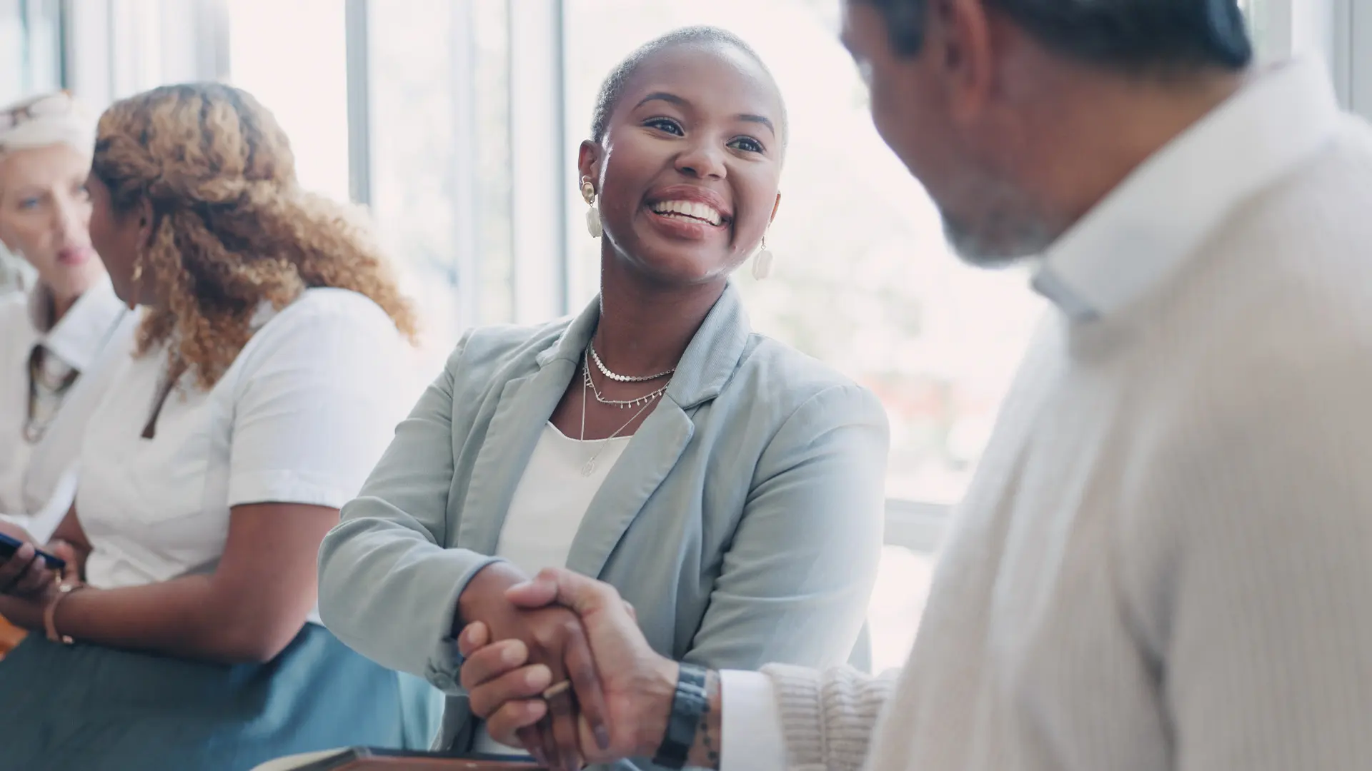 A smiling woman in a business suit shakes hands with a man in a white shirt. Two women converse in the background, creating a warm, professional atmosphere.