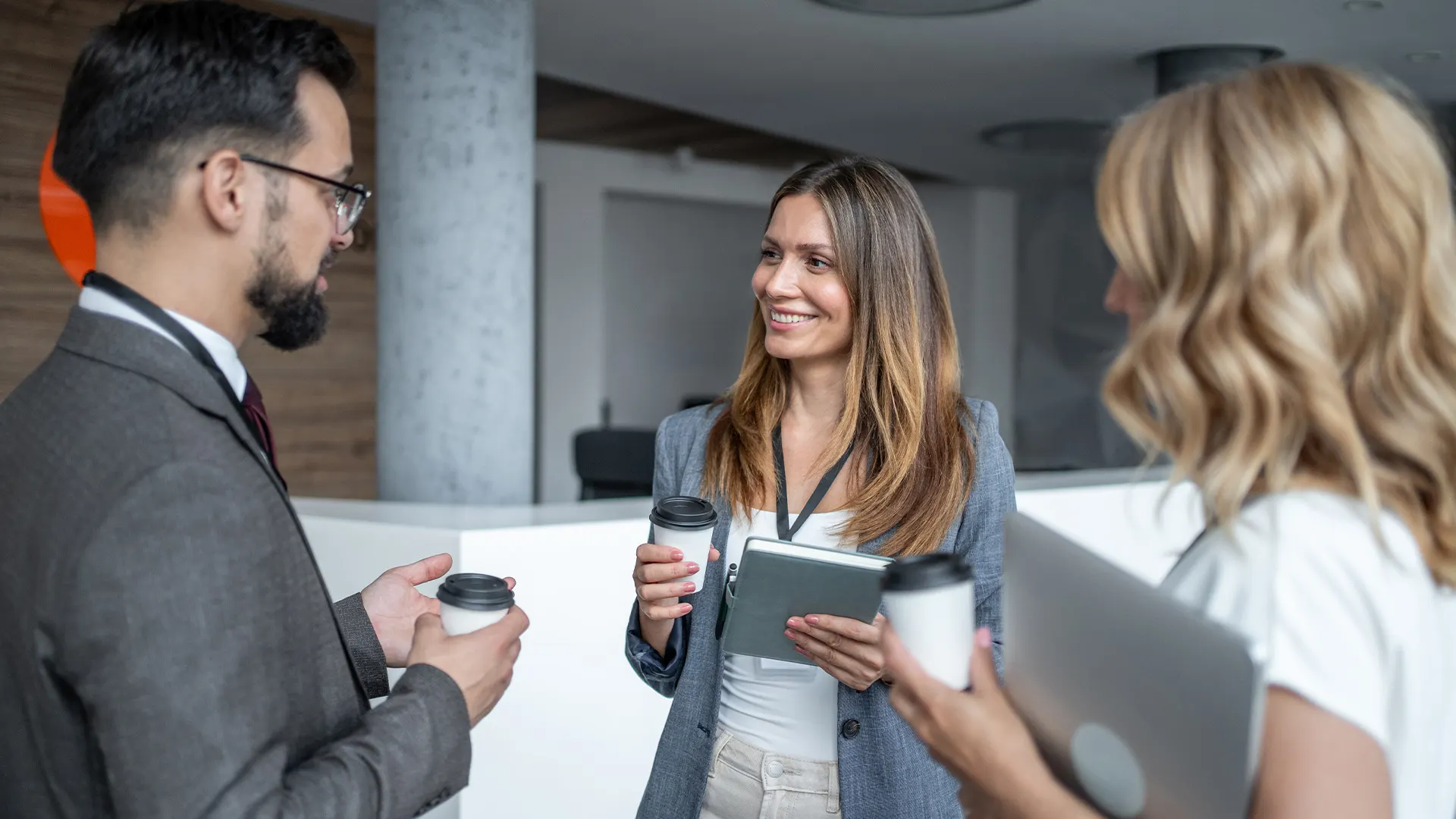 Three professionals standing indoors, holding coffee cups and having a conversation; one person holds a tablet, another holds a laptop.