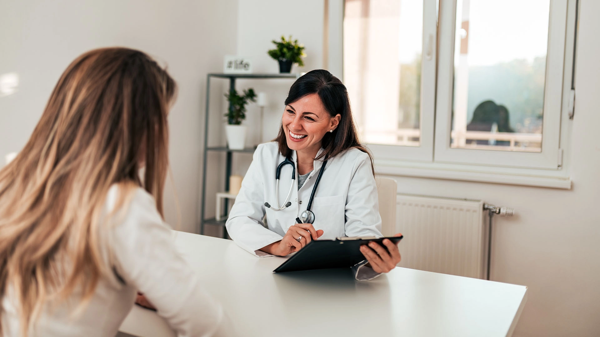 A woman in a white medical coat holds a clipboard and smiles across a white office table at a patient.