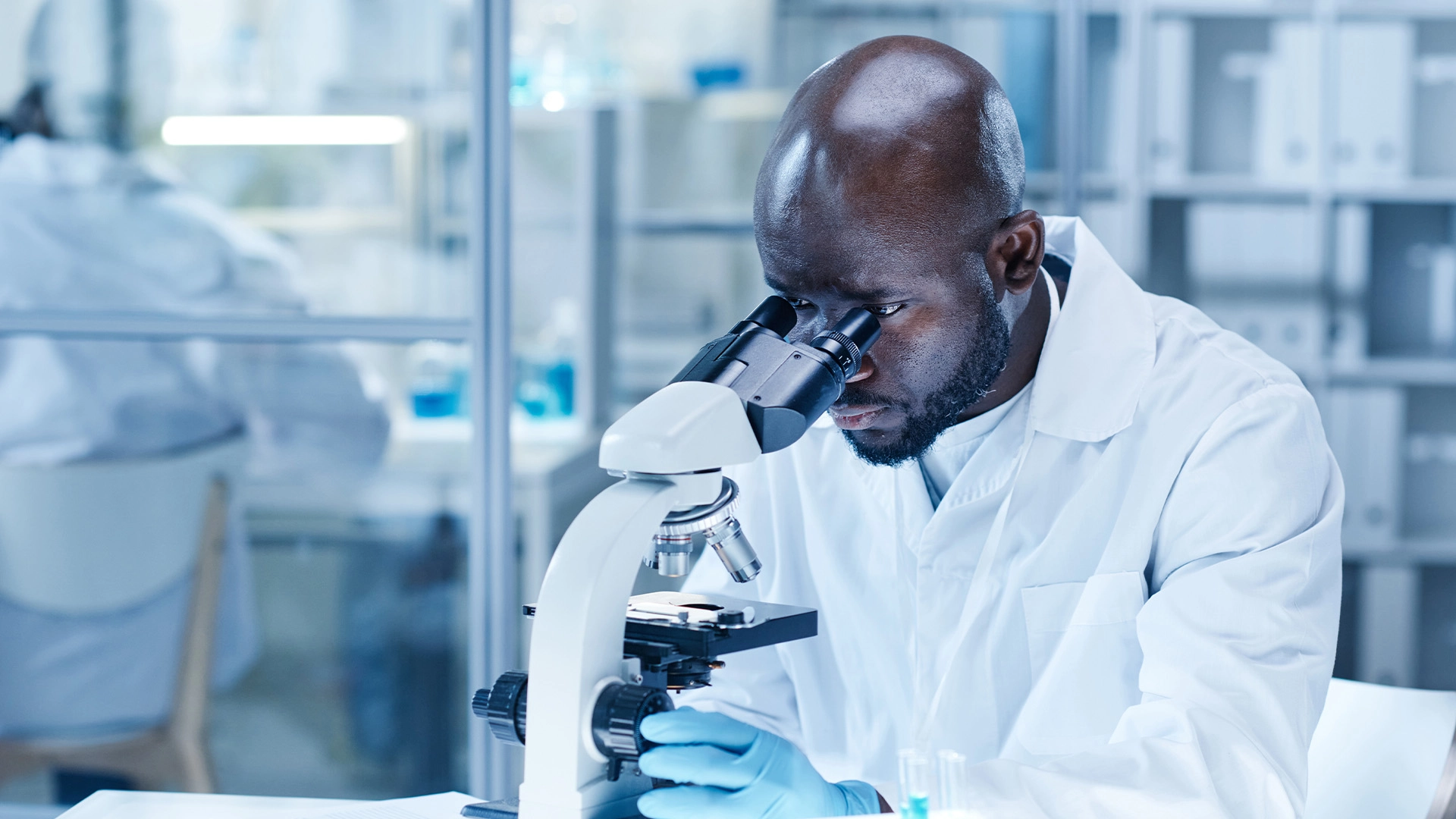 A male scientist wearing a labcoat and nitrile gloves looks trough a microscope lens at a slide within a modern biotech lab.