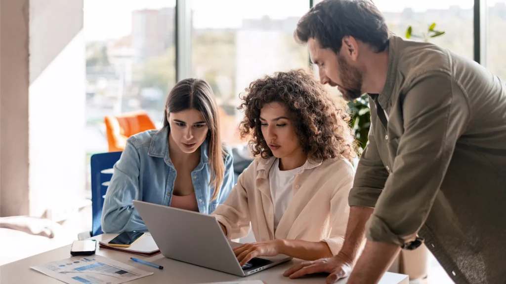 Three colleagues collaborating in a bright office, focusing on a laptop screen. Papers and phones are on the table.