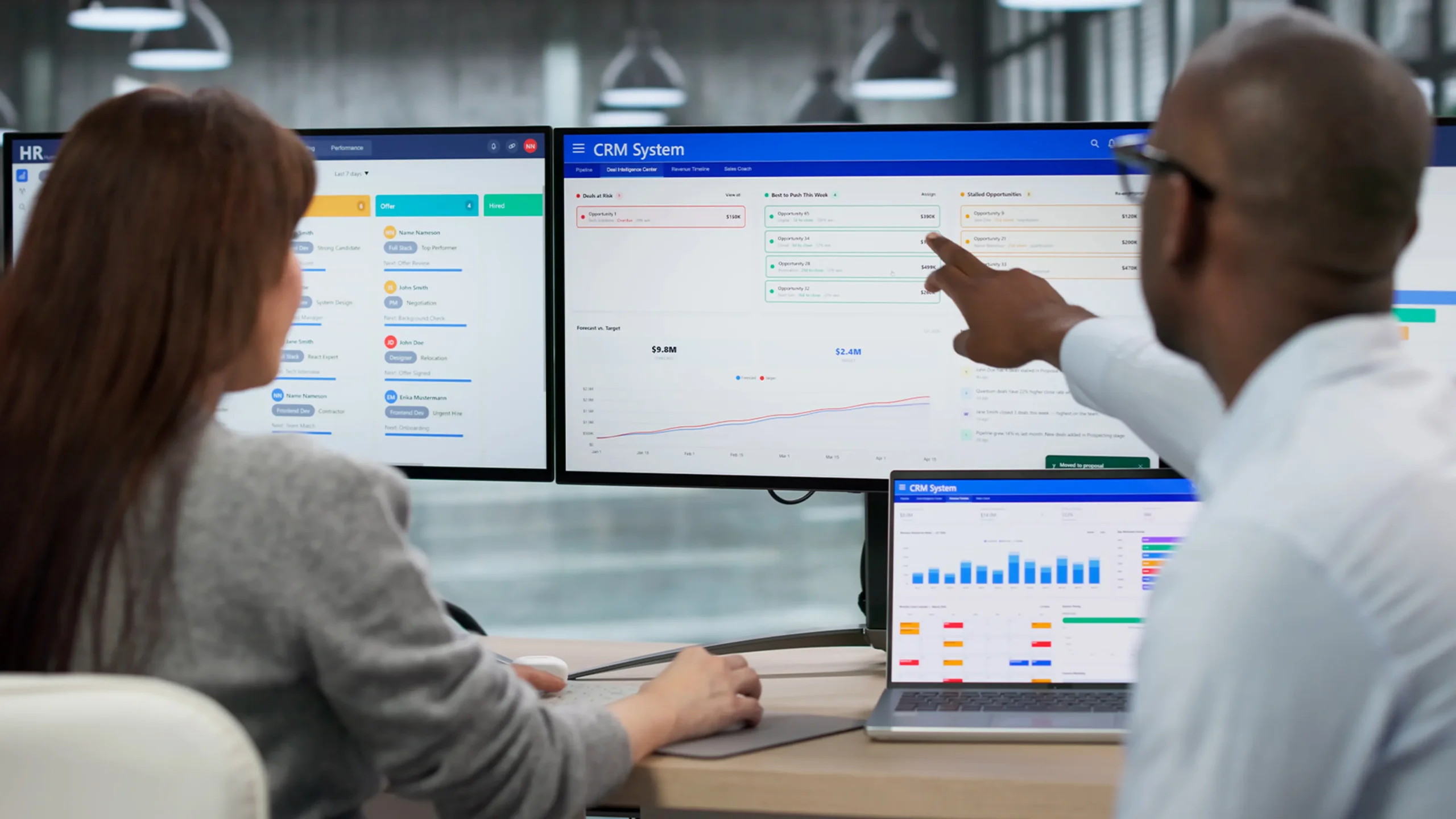 Two people working on large computer monitors. The screen shows a CRM system.