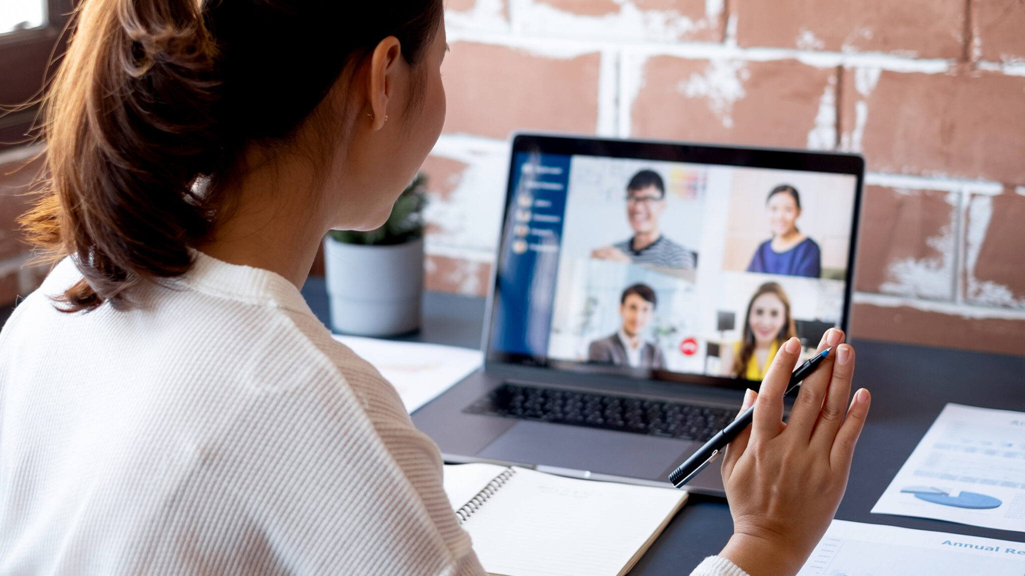 A woman participates in a video conference, looking at a laptop screen displaying four people. She waves while taking notes, conveying an engaged and collaborative tone.